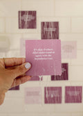 A hand with light purple nail polish holds a wet purple JaxKelly Shower Affirmation™ Card - Boundaries under running water. The card says, “It’s okay if others don’t understand or agree with the boundaries I set.” Blurred cards line a tiled wall behind.