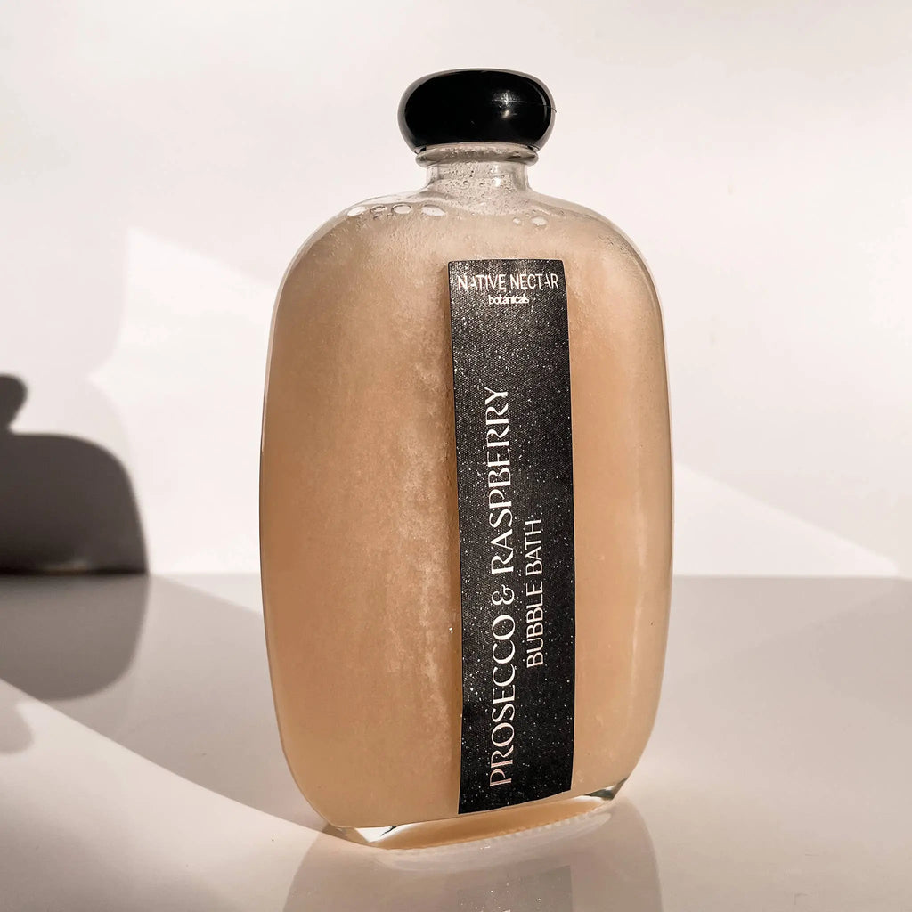 A clear bottle with a black cap contains peach-colored Prosecco & Raspberry Bubble Bath by Native Nectar Botanicals, resting on a white surface with soft shadows.