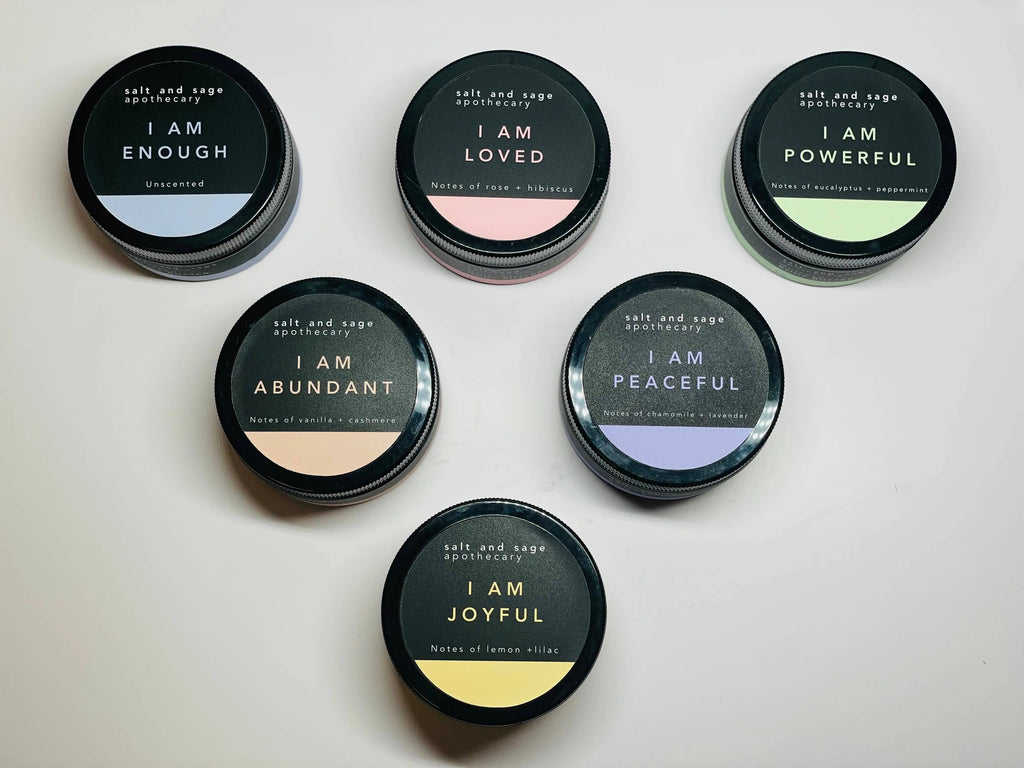 Six black jars from Salt & Sage Apothecary, each labeled with affirmations like I AM ENOUGH and I AM LOVED, hold the I AM Abundant Whipped Organic Body Butter—ideal for luxurious self-care with organic skincare.