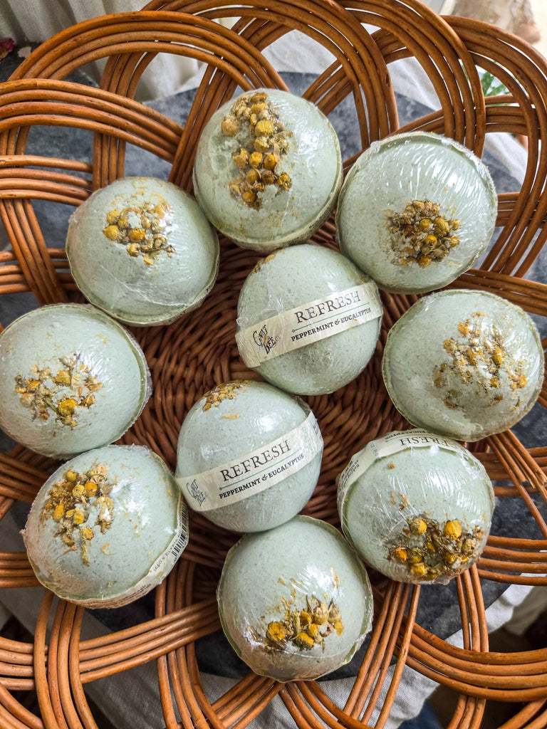 Ten light green Gnat and Bee Refresh | Natural Bath Bombs with dried chamomile flowers rest on a wicker tray. Labeled as peppermint & eucalyptus for respiratory support, each is wrapped in clear plastic.