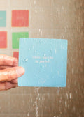 A hand holds a blue JaxKelly Shower Affirmation™ Card for Teens reading “I don’t have to be perfect,” with water droplets visible as the card is used under running shower water against colorful tiled walls.