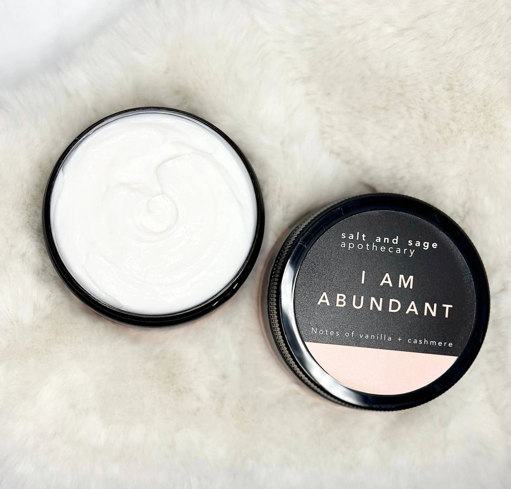 An open jar of Salt & Sage Apothecarys I AM Abundant Whipped Organic Body Butter rests on a soft white surface, the lid beside it reading “Notes of vanilla + cashmere,” inviting you to indulge in luxurious self-care.