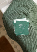 JaxKelly Shower Affirmation™ Cards – Abundance sit atop a green towel, with the top waterproof card stating, “I attract wealth and stability by being my authentic self.” Water droplets add to this inspiring scene.
