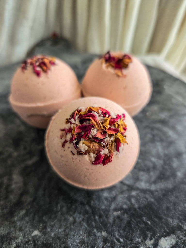 Three Gypsy Rose | Natural Bath Bombs by Gnat and Bee, topped with dried flower petals, rest on a dark textured surface beside a soft curtain—perfect for a stylish, soothing bath that helps you relax and ease anxiety.