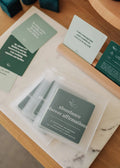 A stack of JaxKelly Shower Affirmation™ Cards - Abundance in teal waterproof design sits on a marble tray, with additional uplifting cards displayed upright behind them on a wooden stand.