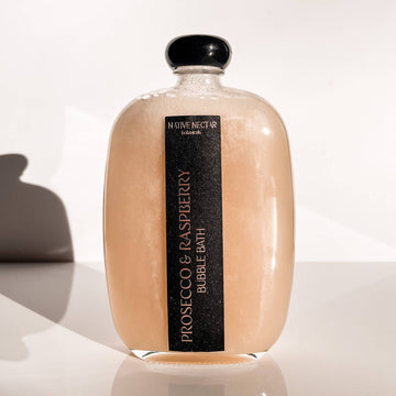 A clear glass bottle of Native Nectar Botanicals Prosecco & Raspberry Bubble Bath, a natural and hydrating formula, rests on a white surface with a soft shadow to the left.