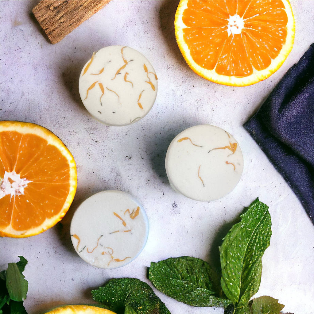 Three round, white Shower Steamers with orange flecks by Woodsy Homelife are displayed on a light surface, surrounded by orange halves, fresh mint, and a cinnamon stick—ideal for a spa-like experience with invigorating essential oil scents.