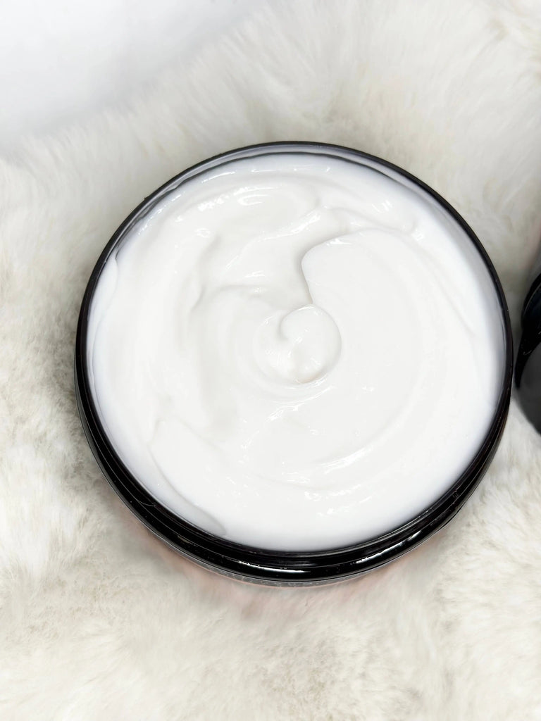An open container of Salt & Sage Apothecary’s I AM Abundant Whipped Organic Body Butter sits on a plush white surface, its creamy, glossy swirl inviting you to indulge in luxurious self-care.