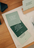 Three green waterproof Shower Affirmation™ Cards - Abundance by JaxKelly, featuring white motivational messages, rest on a marble tray atop a wooden desk with a card holder displaying another uplifting affirmation.