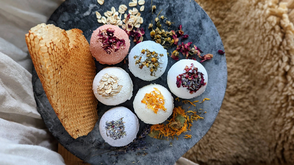 A slate plate displays six Gypsy Rose Natural Bath Bombs by Gnat and Bee, featuring soothing pink grapefruit and dried botanicals, surrounded by petals, oats, and honeycomb for a cozy, anxiety-reducing arrangement.