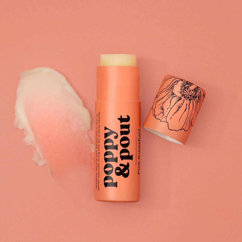 A coral Pink Grapefruit Lip Balm by Poppy & Pout with the cap off sits beside a smear of balm on a matching pink background. The eco-friendly cap displays a delicate poppy flower illustration.