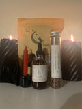 The Divine Love Spell Kit by Luna & Co. Soapery, featuring a dropper bottle, spell jar, red and black candles, pouch, and branded bag, is arranged on a white surface.