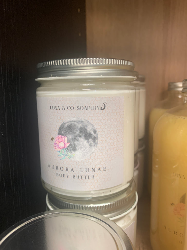 A jar of Luna & Co. Soaperys Aurora Lunae Body Butter, made with shea butter and adorned with a moon and pink flower label, sits on a shelf beside other jars and bottles.