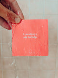 A hand holds a red JaxKelly Shower Affirmation™ Card - Teens under running water. The waterproof card, reading “I can always ask for help” with a smiley face, is covered in droplets against a tiled wall.