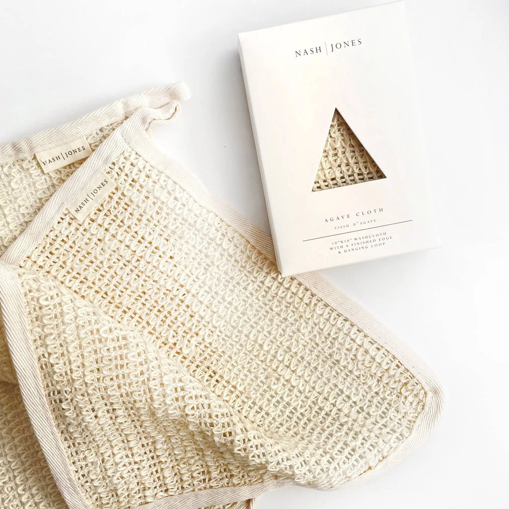 A cream-colored, textured Nash and Jones Agave Wash Cloth is displayed beside its packaging, which features a triangular cutout revealing the Natural Agave Wash Cloth inside.