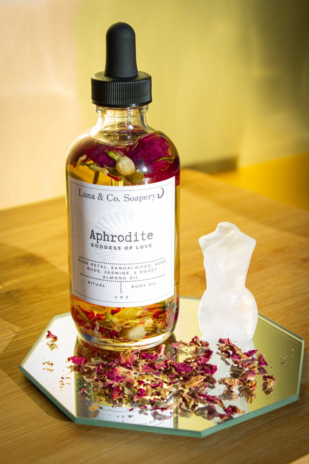 A glass bottle of Luna & Co. Soaperys Aphrodite Body Oil sits on a mirror with dried petals, next to a white torso-shaped candle, all arranged on a wooden surface bathed in warm light.