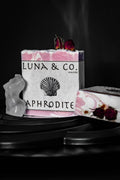 A pink and white marbled bar from Luna & Co. Soaperys Aphrodite Glow Bundle, topped with dried roses and featuring a shell illustration—perfect for self-care rituals, shown on a dark surface with other rose-topped soaps.