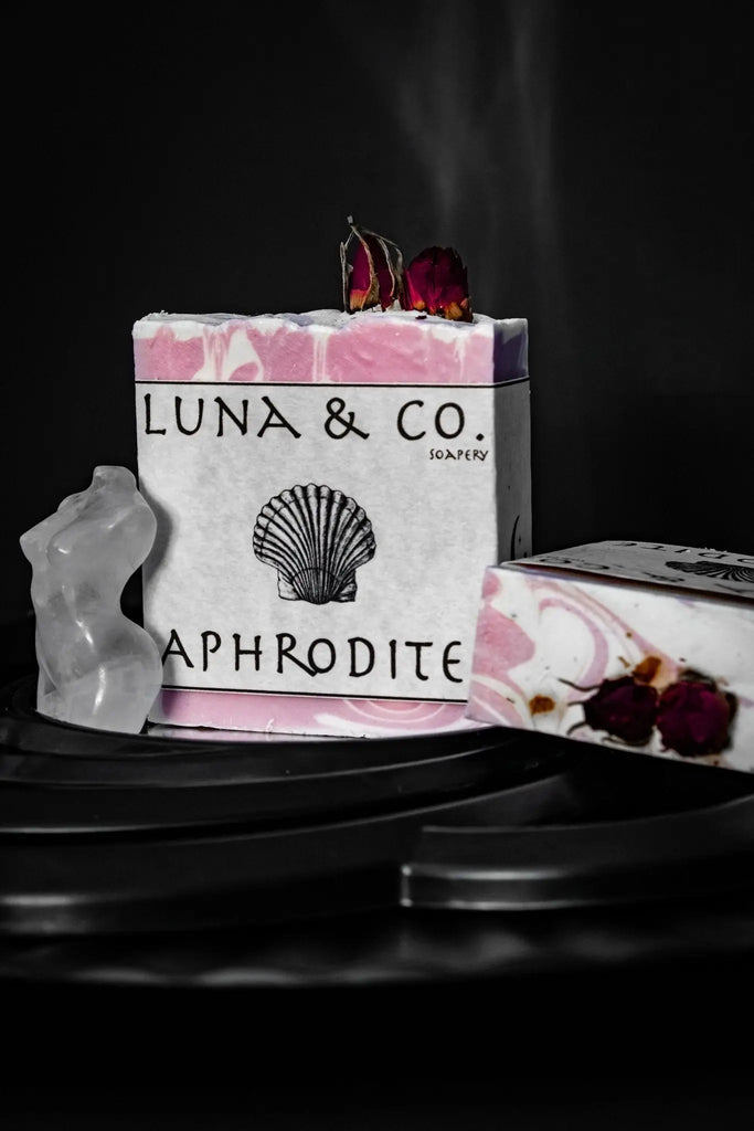 A pink and white marbled bar from Luna & Co. Soaperys Aphrodite Glow Bundle, topped with dried roses and featuring a shell illustration—perfect for self-care rituals, shown on a dark surface with other rose-topped soaps.