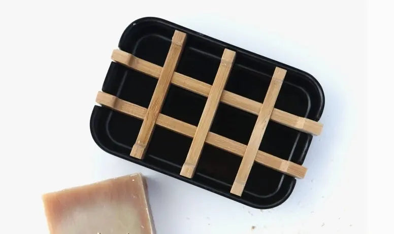 A black soap dish with a wooden grid rests beside a brown bar of soap on a white surface, adding elegance to your ritual and perfectly complementing the Aphrodite Glow Bundle by Luna & Co. Soapery.