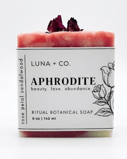 A 5 oz bar of Luna + Co. Aphrodite Ritual Botanical Soap, featuring a pink and white marbled soap bar with dried rose petals on top, wrapped in a white paper label with black text and rose line art.