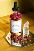 A glass dropper bottle of Luna & Co. Soaperys Artemis Body Oil with dried rose petals rests on a mirror beside scattered petals and a clear quartz crystal, all arranged on a wooden surface.