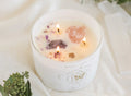 The Air Elements Candle 12oz by THE MINDFUL COLLECTIVE is a white crystal candle in a glass container with three flames, crystals, gemstones, and an astrological label with a butterfly. Greenery and a white selenite wand rest beside this Zodiac Candle.