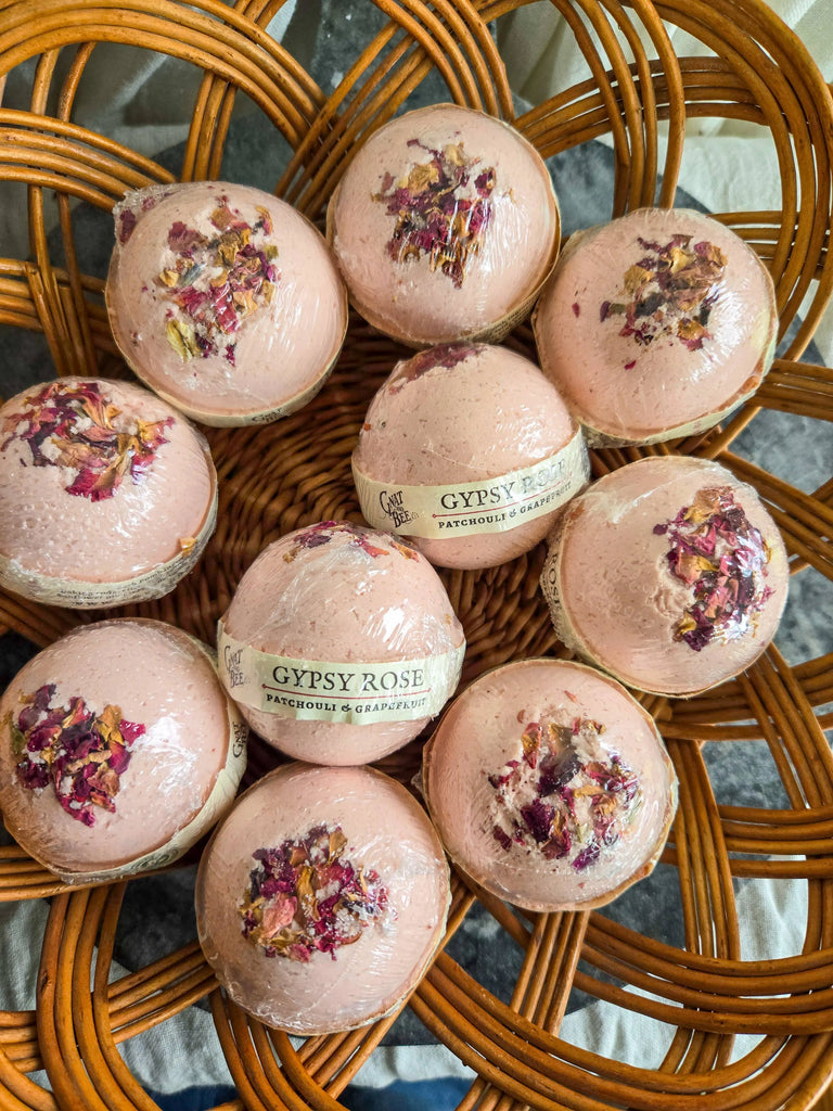 Nine pink Gypsy Rose | Natural Bath Bombs by Gnat and Bee, topped with dried rose petals and scented with pink grapefruit, sit in a wicker basket. Two are labeled for their anxiety-reducing properties during each soothing soak.