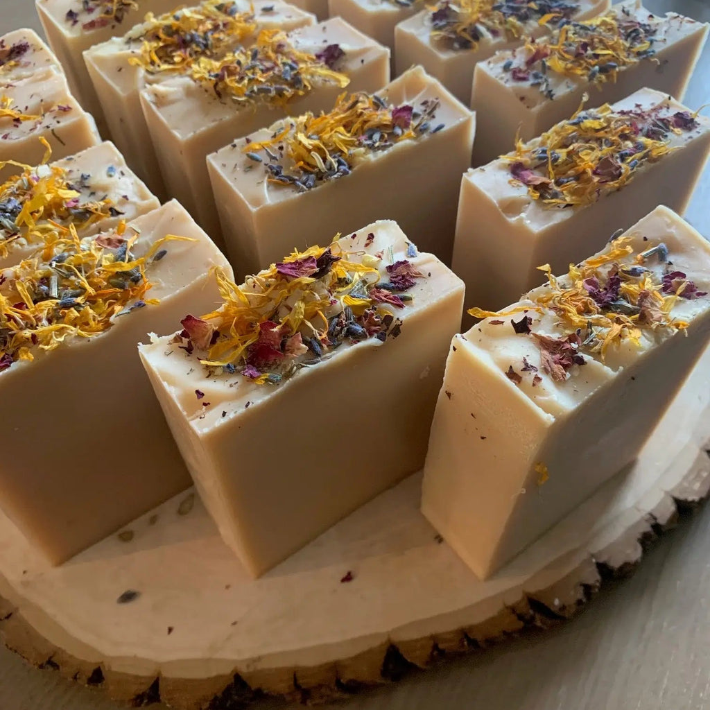 The Bealtaine Sabbat Bar by Luna & Co. Soapery features natural ingredients and is topped with dried flower petals and herbs, creating a beautifully decorative, artisanal soap displayed on a wooden slab.