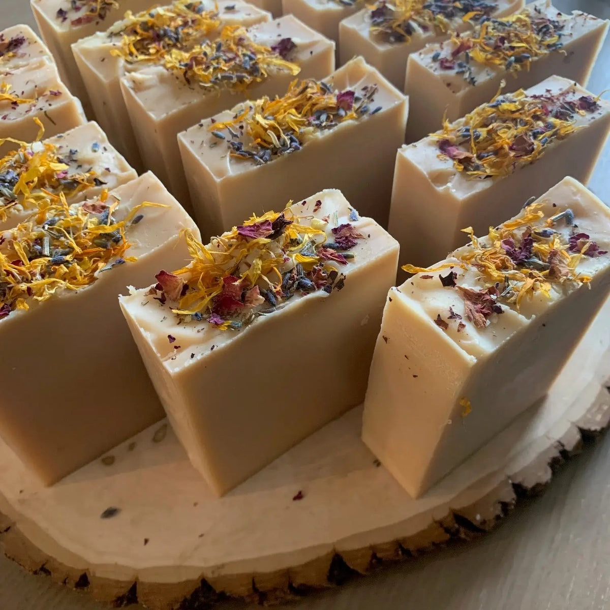 The Bealtaine Sabbat Bar by Luna & Co. Soapery features natural ingredients and is topped with dried flower petals and herbs, creating a beautifully decorative, artisanal soap displayed on a wooden slab.