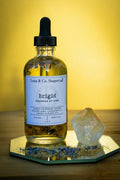 A glass bottle of Luna & Co. Soaperys Brigid Body Oil rests on a mirror with dried flowers and a crystal nearby, set against a yellow background.