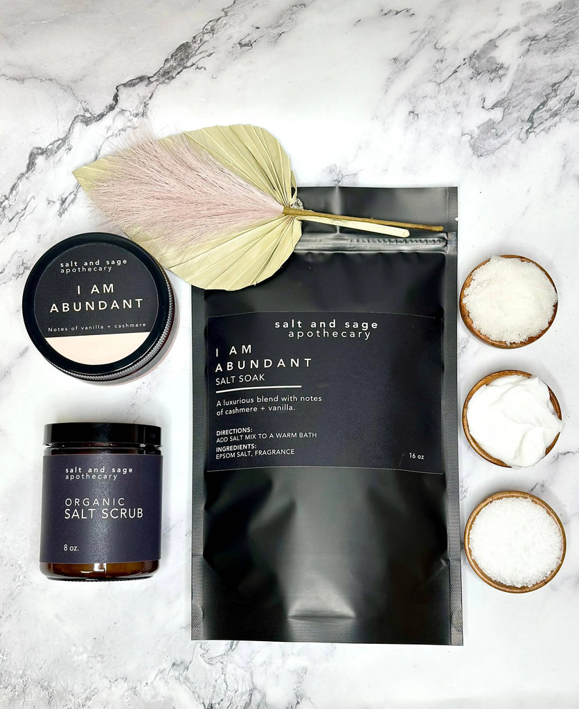 A flat lay of Salt & Sage Apothecary’s I AM Abundant Whipped Organic Body Butter with salt soak, organic salt scrub, bowls of salt and powder, and a dried palm leaf on marble—ideal for luxurious self-care.