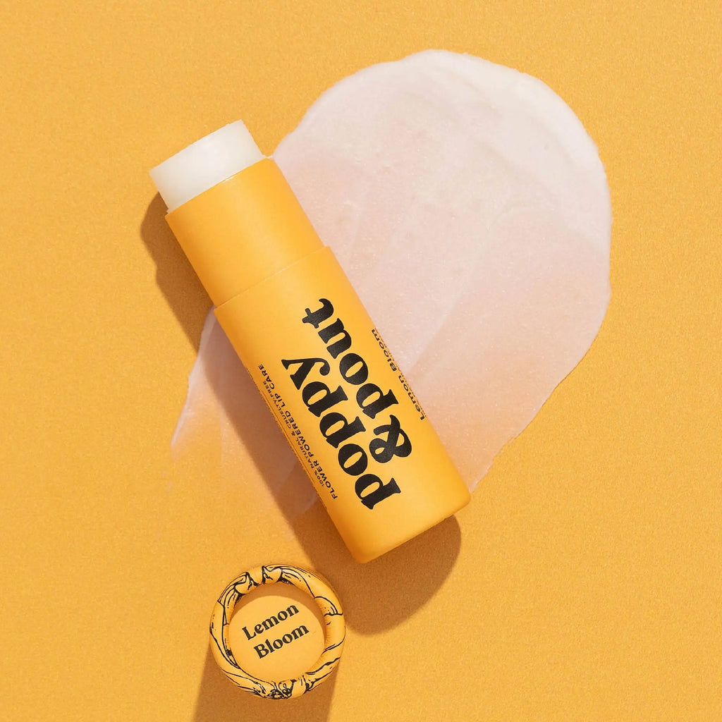 A yellow tube of Poppy & Pout Lemon Bloom Lip Balm rests on a textured yellow background with a swipe of natural balm behind it. The round cap, nearby, is labeled with Lemon Bloom.