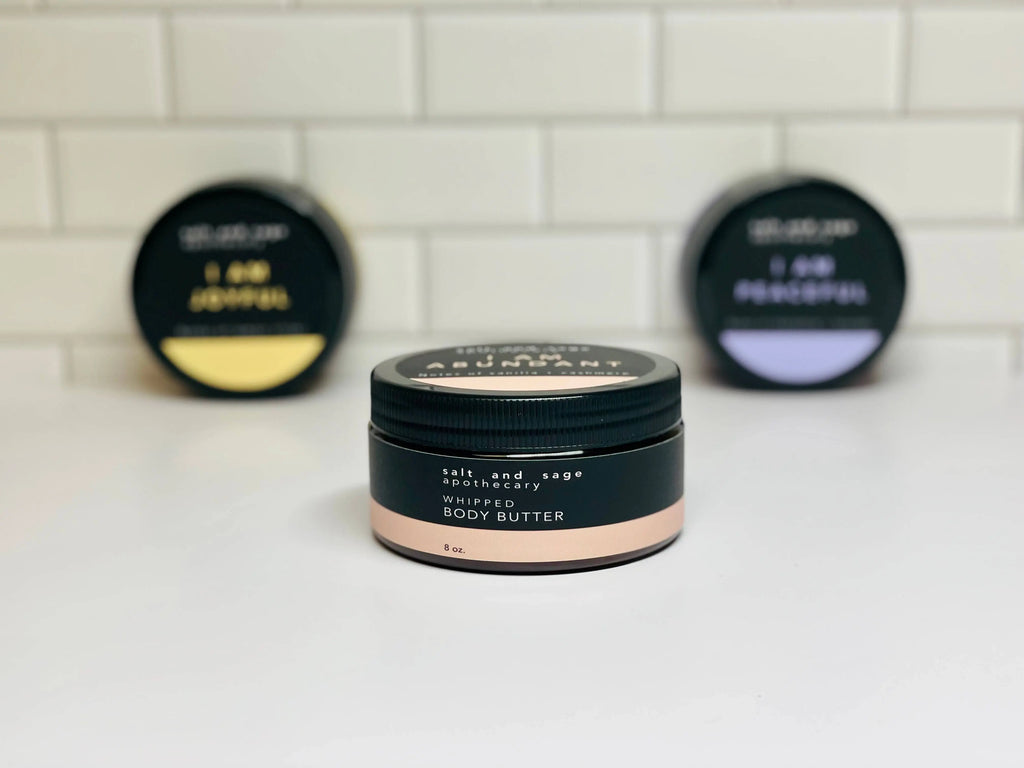 A small jar of Salt & Sage Apothecary I AM Abundant Whipped Organic Body Butter sits in focus on a white surface, with two blurred jars labeled I AM Joyful and I AM Peaceful behind it, suggesting a luxurious self-care experience.
