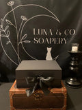 A black gift box with a satin ribbon rests on a brown leather trunk. Behind it, a black backdrop displays Luna & Co. Soapery with celestial graphics. The Celestial Gift Set includes Planetary Soaps and a candle on the right.
