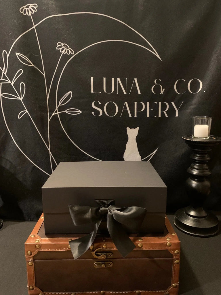 A black gift box with a satin ribbon rests on a brown leather trunk. Behind it, a black backdrop displays Luna & Co. Soapery with celestial graphics. The Celestial Gift Set includes Planetary Soaps and a candle on the right.