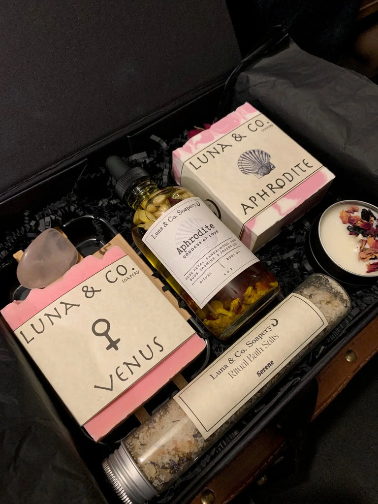 The Celestial Gift Set by Luna & Co. Soapery includes Venus and Aphrodite soaps, floral bath oil, a dried petal candle, soothing bath salts, and a crystal—all nestled in black shredded paper.