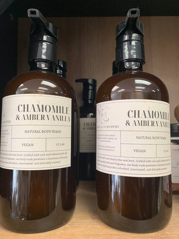 Two brown bottles of Chamomile body wash by Luna & Co. Soapery with black pumps are shown on a shelf. The vegan label is visible.