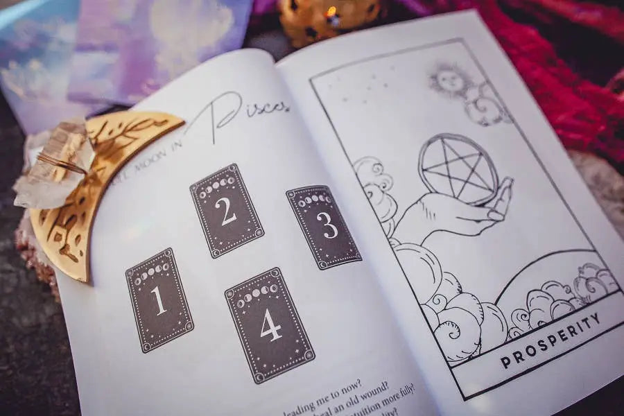 The Cosmic Guidance Book by Bella deLuna is open to a four-card tarot spread guide featuring a hand holding a pentacle labeled “Prosperity,” with crystals and a wooden crescent moon atop the pages for a celestial, insightful reading.