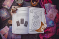 The Cosmic Guidance Book by Bella deLuna lies open, showcasing a lion tarot card and Pluto Retrograde page, surrounded by tarot spreads, crystals, candles, a wooden star, and a crescent moon—crafting a celestial scene on the dark surface.