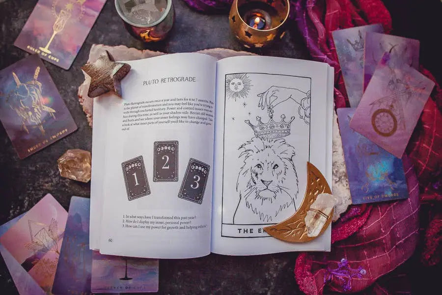 The Cosmic Guidance Book by Bella deLuna lies open, showcasing a lion tarot card and Pluto Retrograde page, surrounded by tarot spreads, crystals, candles, a wooden star, and a crescent moon—crafting a celestial scene on the dark surface.