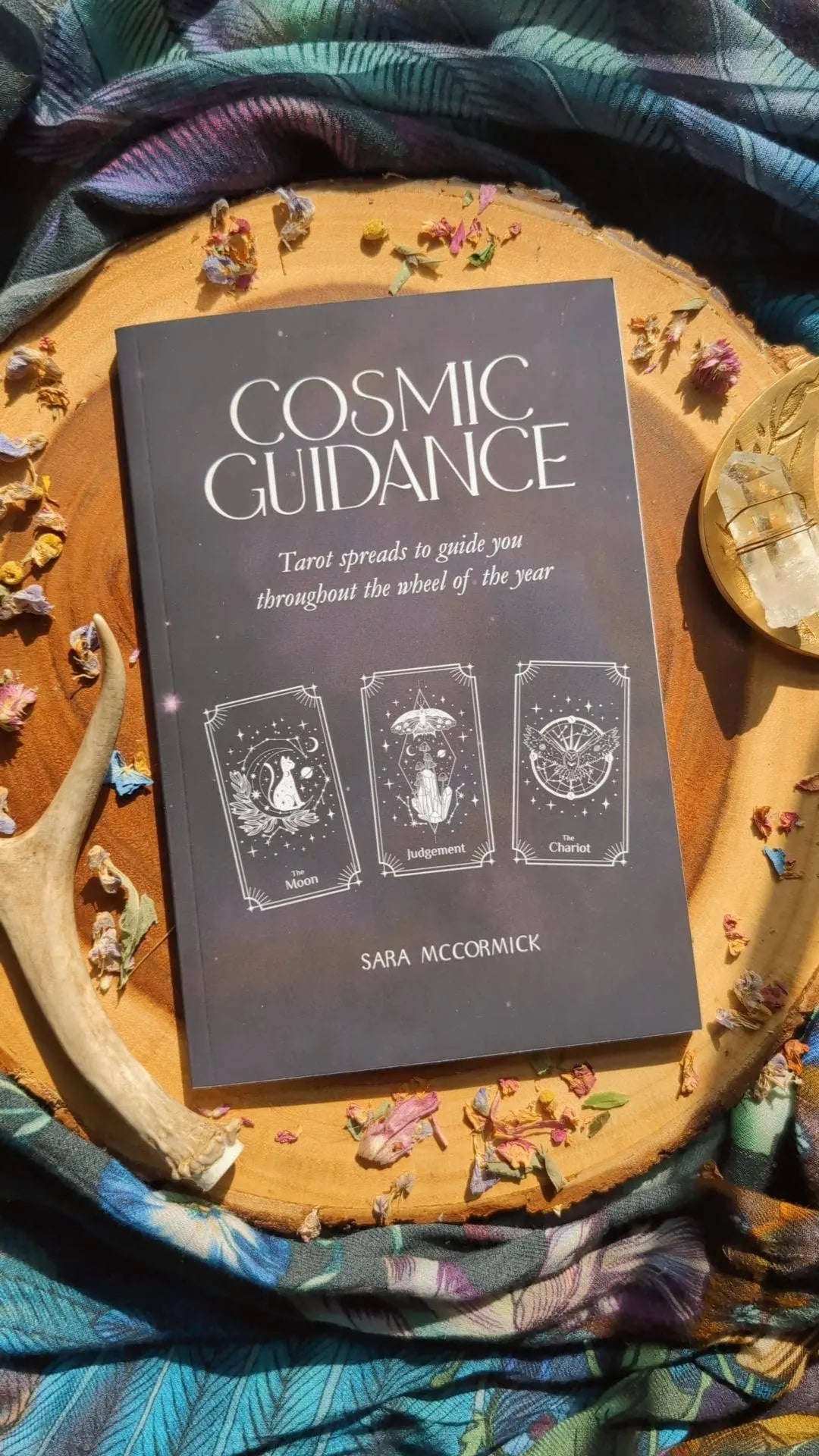 The Cosmic Guidance Book by Bella deLuna rests on a wooden tray with colorful fabric, dried flowers, an antler, and a crystal. Its cover shows three tarot cards—Moon, Judgment, and Chariot—for mystical inspiration.