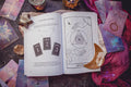 The Cosmic Guidance Book by Bella deLuna features tarot card illustrations, a moon phase chart, and celestial roadmap motifs. Displayed with tarot cards, crystals, candles, a crescent moon ornament, and purple cloth on a wooden surface.