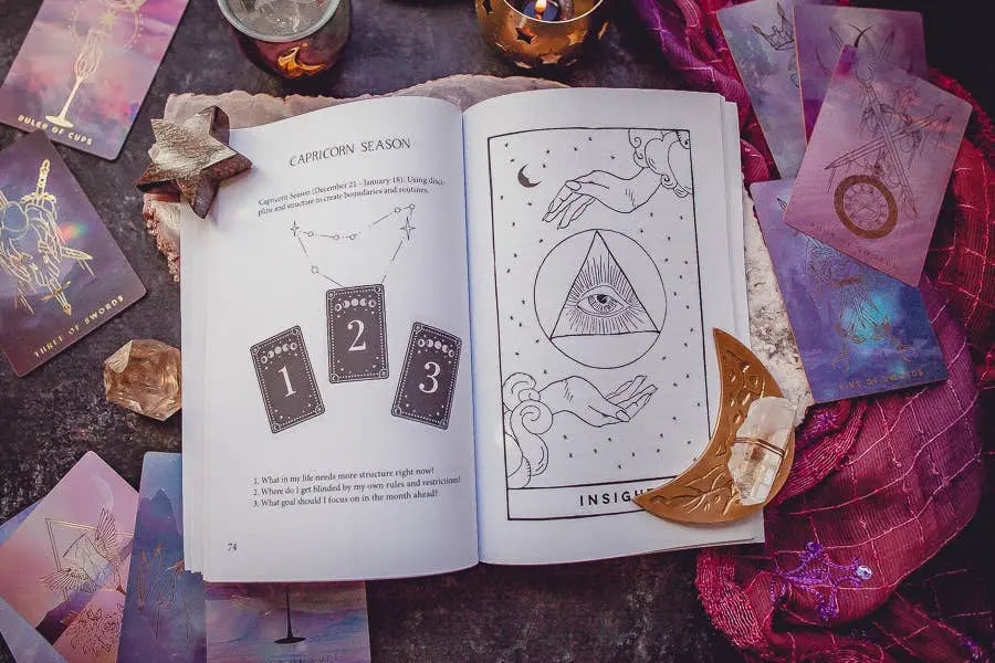 The Cosmic Guidance Book by Bella deLuna features tarot card illustrations, a moon phase chart, and celestial roadmap motifs. Displayed with tarot cards, crystals, candles, a crescent moon ornament, and purple cloth on a wooden surface.