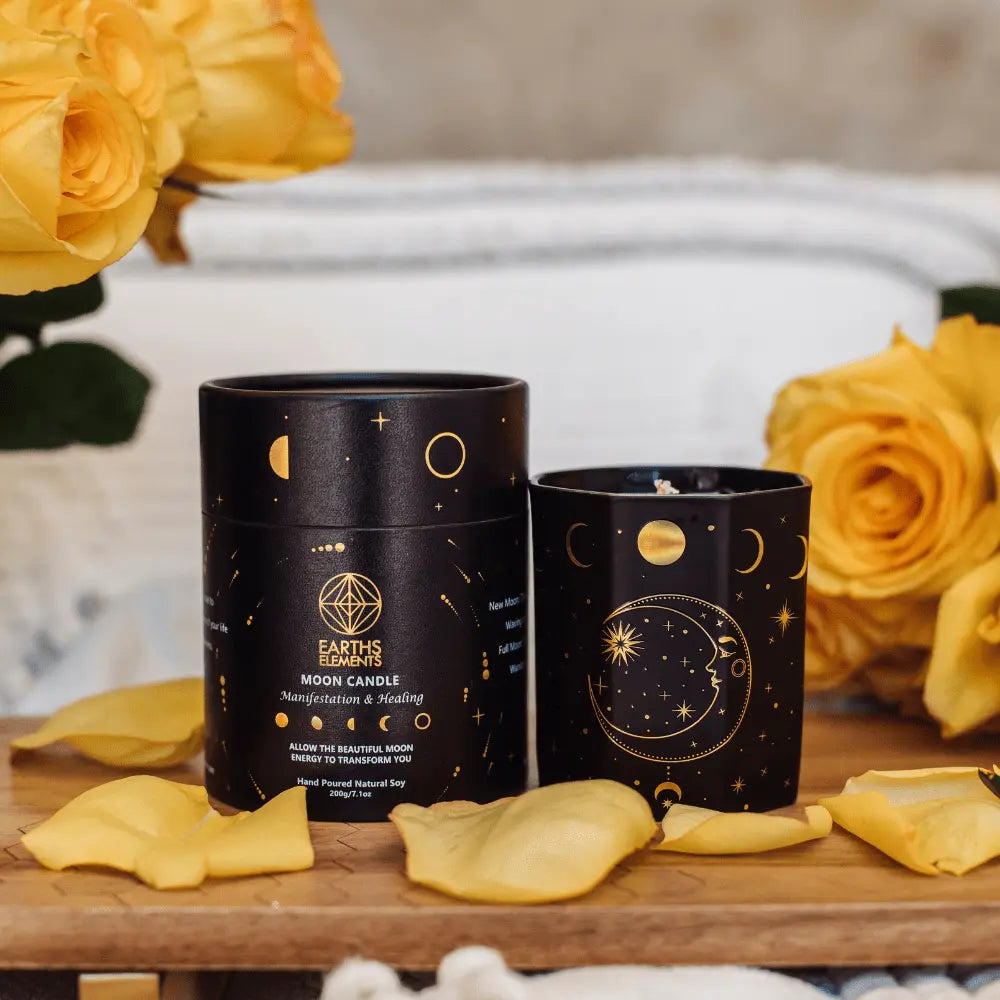 A black jar adorned with moon phase designs, from Earths Elements Wholesales Crystal Candle, Moon Phase - pack of 6, sits next to its matching box amid yellow roses and petals on a white surface.