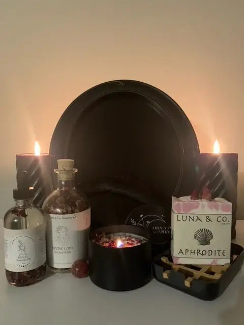 The Divine Love Ritual Kit by Luna & Co. Soapery features two candles, a black mirror, incense, a gemstone, and labeled bottles—arranged on a flat surface to inspire self-love.