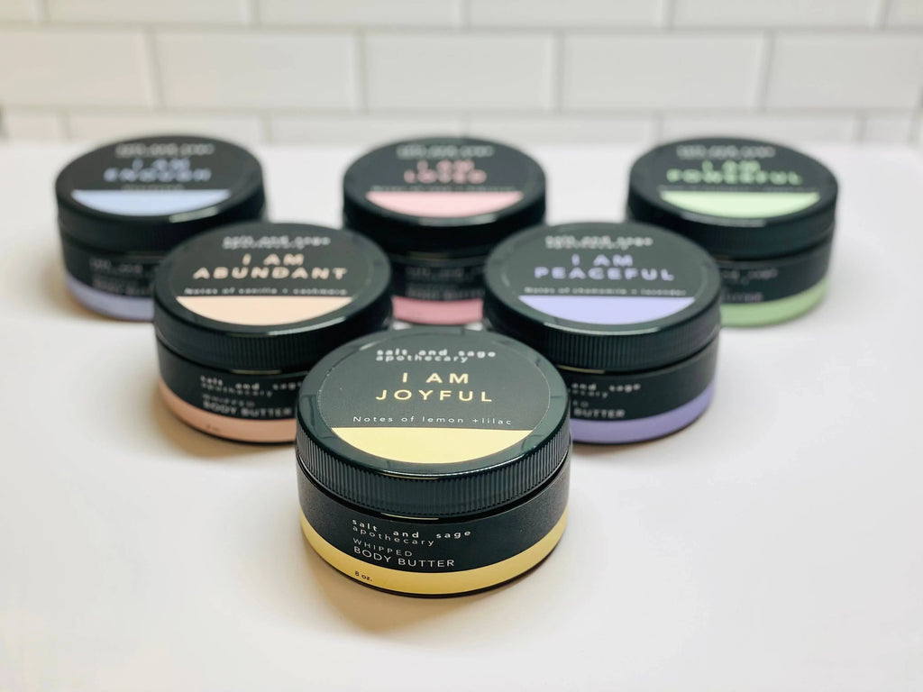 Seven jars of Salt & Sage Apothecarys I AM Abundant Whipped Organic Body Butter with black lids are displayed on a white surface, each featuring pastel labels and uplifting affirmations—ideal for an organic skincare routine.
