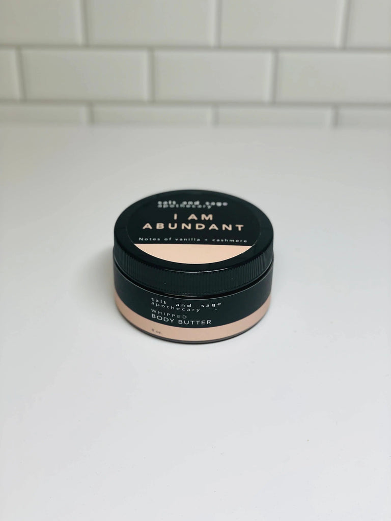 A jar of Salt & Sage Apothecary’s I AM Abundant Whipped Organic Body Butter sits on a white surface, featuring a black lid and light base. This luxurious self-care treat envelops you in soothing scents of vanilla and cashmere.