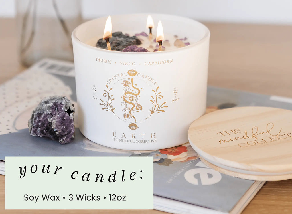 A white Earth Elements Candle 12oz by THE MINDFUL COLLECTIVE with crystals on top sits on magazines next to a wooden lid and amethyst stones. Overlay: Soy Wax • 3 Wicks • 12oz. Perfect for Taurus, Virgo, or Capricorn.