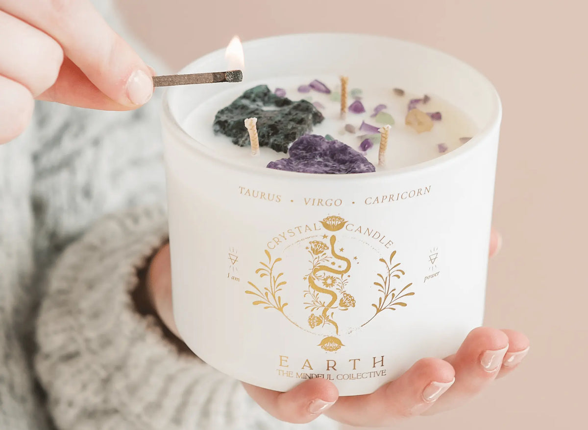 A person lights THE MINDFUL COLLECTIVE Earth Elements Candle 12oz. The candle has purple and green crystals inside, with gold Taurus, Virgo, and Capricorn zodiac symbols on the jar.