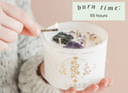 A person lights the Earth Elements Candle 12oz by THE MINDFUL COLLECTIVE, featuring stones inside. The label lists Taurus, Virgo, Capricorn. Burn time: 55 hours—a grounding candle perfect for Earth Signs.